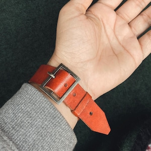 May include: A burnt orange leather watch strap with a large, square, silver-colored buckle. The strap is secured on a wrist, showing the texture and color of the leather. The watch band is approximately 1 inch wide.