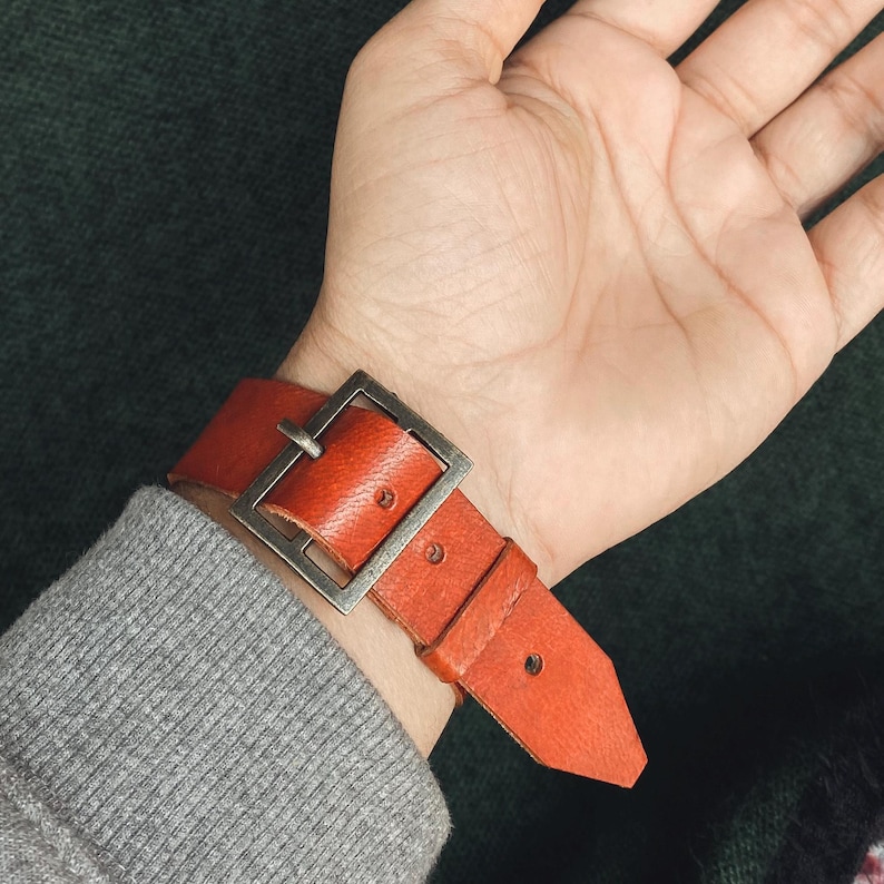 May include: A burnt orange leather watch strap with a large, square, silver-colored buckle. The strap is secured on a wrist, showing the texture and color of the leather. The watch band is approximately 1 inch wide.