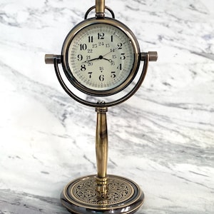 May include: Antique brass-colored desk clock with a round face and a stand. The clock face has black numerals and hands, with a second set of numbers around the edge. The clock is supported by a decorative base.