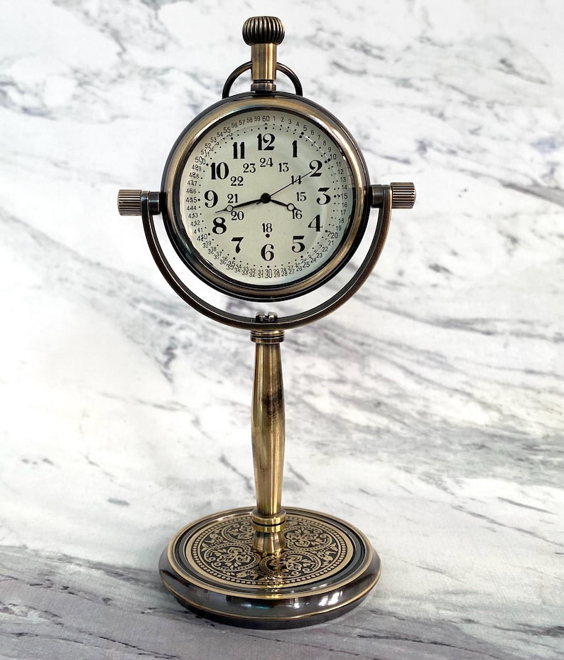 May include: Antique brass-colored desk clock with a round face and a stand. The clock face has black numerals and hands, with a second set of numbers around the edge. The clock is supported by a decorative base.