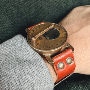 May include: A vintage brass sundial watch with a red leather strap. The watch face features etched Roman numerals and a central gnomon. The strap is secured with two metal rivets. A unique, antique-style timepiece.