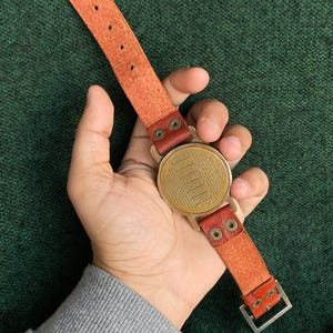 May include: A vintage-style watch with a round, brass-colored face and a tan leather strap. The watch face has a detailed, engraved design. The strap features a buckle and is attached with brass-colored rivets. The watch is held against a green background.