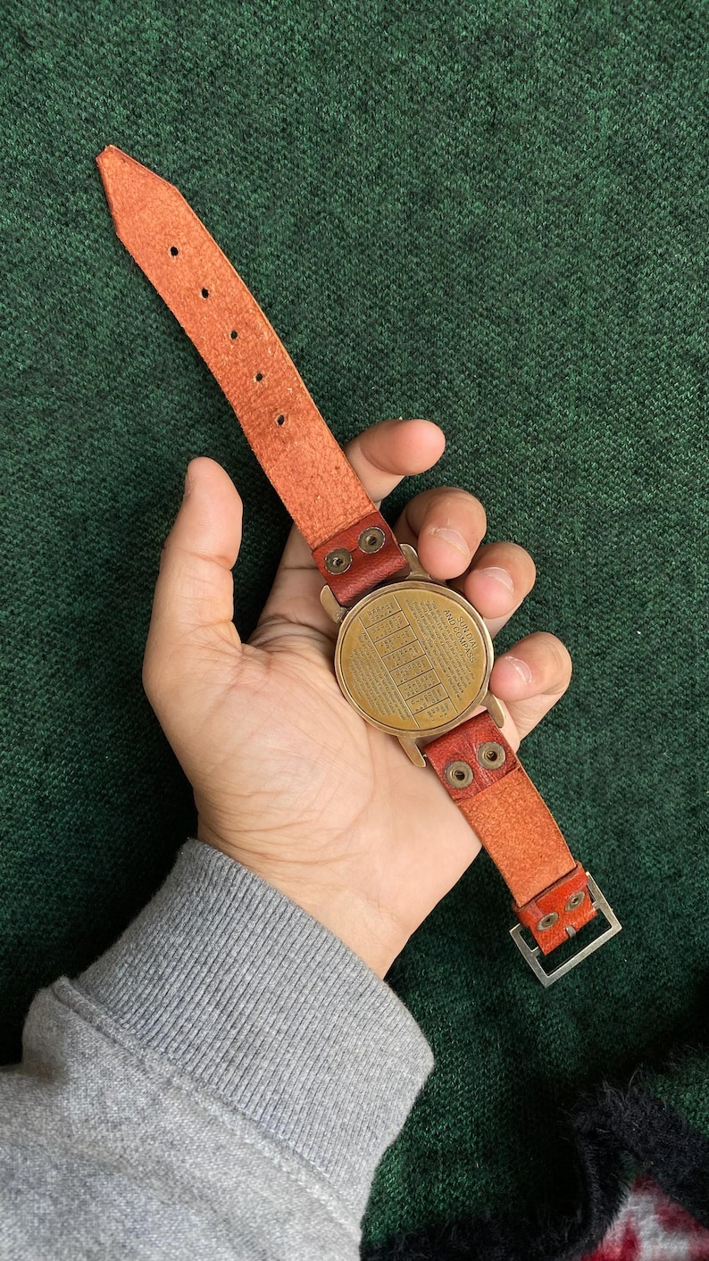 May include: A vintage-style watch with a round, brass-colored face and a tan leather strap. The watch face has a detailed, engraved design. The strap features a buckle and is attached with brass-colored rivets. The watch is held against a green background.