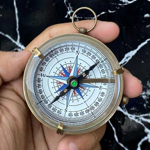 Peut inclure: Une boussole en laiton vintage avec un cadran blanc orné d'une rose des vents bleue, rouge et verte. La boussole a un couvercle en verre transparent et un anneau pour l'accrocher. La boussole est tenue dans une main.