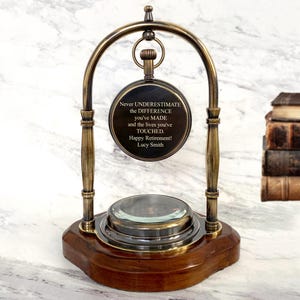 Könnte beinhalten: Ein Schreibtischornament im Vintage-Stil mit Holzsockel und messingfarbenem Metallrahmen. Das Ornament verfügt über eine Lupe und eine runde Plakette mit dem Text "Never UNDERESTIMATE the DIFFERENCE you've MADE... Happy Retirement! Lucy Smith."