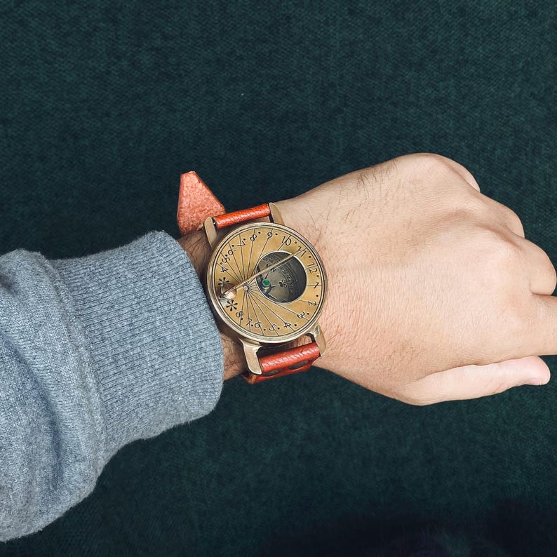 May include: A brass-colored sundial watch with a brown leather band is worn on a wrist. The watch face features a circular design with markings and a small, green element. The watch is set against a dark green background.
