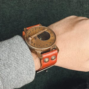 May include: A vintage-style brass sundial watch with a brown leather strap. The watch face features etched Roman numerals and a central gnomon. The leather strap is secured with two brass rivets.