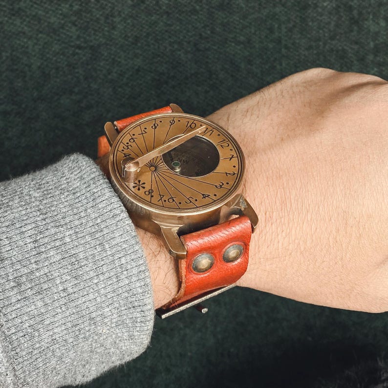 May include: A vintage-style brass sundial watch with a brown leather strap. The watch face features etched Roman numerals and a central gnomon. The leather strap is secured with two brass rivets.