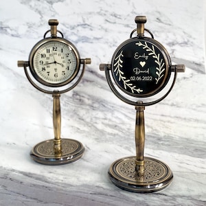 May include: Two antique-style desk clocks with bronze-colored metal frames. One clock displays a traditional clock face with black numerals on a white background. The other clock has a black face with white text that reads "Emily David 02.06.2022".