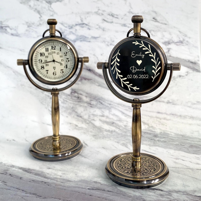 May include: Two antique-style desk clocks with bronze-colored metal frames. One clock displays a traditional clock face with black numerals on a white background. The other clock has a black face with white text that reads "Emily David 02.06.2022".