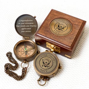May include: A vintage brass compass with a chain, open to reveal a message. A wooden box with a brass emblem and a second compass are also present. The compasses and box feature the United States Navy emblem and text.