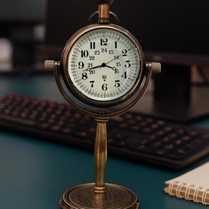 May include: A vintage-style brass desk clock with a round face. The clock features a white dial with black numerals and hands, and a second set of numbers around the edge. The clock is supported by a brass stand with a decorative base.