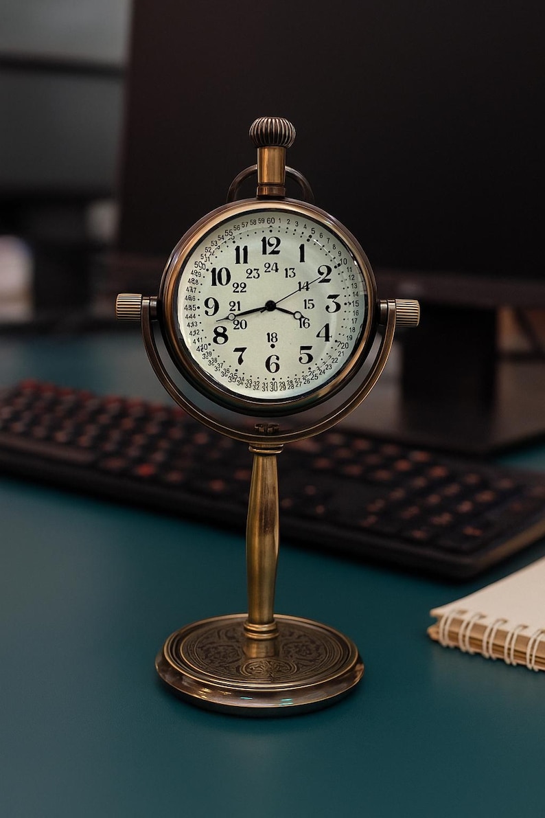 May include: A vintage-style brass desk clock with a round face. The clock features a white dial with black numerals and hands, and a second set of numbers around the edge. The clock is supported by a brass stand with a decorative base.