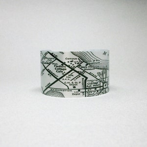 May include: A silver-toned metal cuff bracelet featuring a black printed map design. The map displays street names like Regent St and Washington, along with landmarks such as the State Historical Museum, City Hall, and the University of Wisconsin Boathouse.