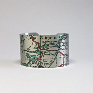 May include: A wide cuff bracelet made of metal featuring a vintage map print. The map displays geographical details including the text Brighton, Rainbow, Paul Smiths, and Upper St. Regis, with red road lines and green shaded areas on a light background.