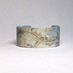 May include: A silver cuff bracelet featuring a topographic map design. The map includes the text "Dana Butte" and "Horse Creek Rapids" with blue river details. The bracelet is approximately 1.5 inches wide.