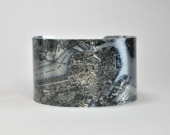 May include: A wide, silver cuff bracelet with a map design in black and white. The map appears to be of a city, with streets and buildings visible. The bracelet is made of a shiny material and has a smooth finish.