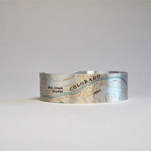 May include: A silver metal cuff bracelet with a topographic map of Colorado. The map shows a river, mountains, and the text "COLORADO" and "Salt Creek Rapids".