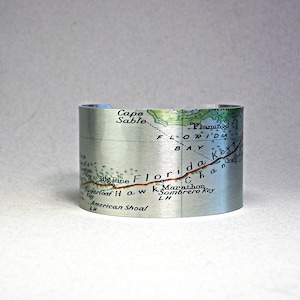 May include: A silver cuff bracelet featuring a map of Florida. The map includes place names like Cape Sable, Flamingo, and Marathon. The bracelet is cylindrical and has a brushed metal finish.