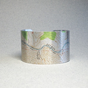 May include: A silver metal cuff bracelet with a topographic map design. The map shows a river, a trail, and the words "Salmon Trail".
