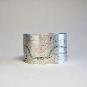 May include: A silver metal cuff bracelet with a map design featuring a river and the text "Green Stillwater Rim".