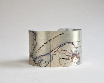May include: A wide silver-toned metal cuff bracelet featuring a detailed vintage map print. The map displays geographical features including Vineyard Haven Harbor, Eastville, and Cottage City, with text labels clearly visible across the surface.