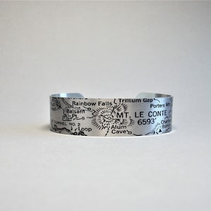 May include: A silver metal bracelet featuring an engraved map of Mount Le Conte in the Great Smoky Mountains National Park. The map includes details such as Rainbow Falls, Trillium Gap, Alum Cave, and Porters Mountain. The elevation of Mount Le Conte is also engraved on the bracelet: 6593'.