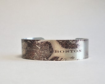 May include: A silver metal cuff bracelet with a vintage map of Boston printed on it. The map is in brown and white and the word "BOSTON" is printed in white on the bracelet.