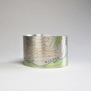 May include: A silver metal cuff bracelet with a topographic map design. The map shows contour lines and the words "MIDDLE FORK" and "FOREST EDGE".