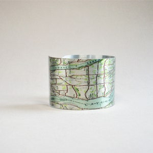 May include: A wide metal cuff bracelet featuring a printed vintage map design. The map displays geographical details including Owasco Lake and Cayuga Lake, with green, tan, and black ink lines visible on the metallic silver surface of the band.