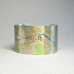 May include: A silver metal bracelet with a topographic map design. The map shows a river, hills, and the text "Middle Fork Redbank River National Forest".