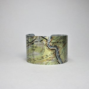 May include: A silver metal cuff bracelet with a topographic map design. The map shows the Delaware Water Gap area with the text "Delaware Water Gap", "Foxtown Gap", "Colledonia", "Mt Minis", and "Slateford".
