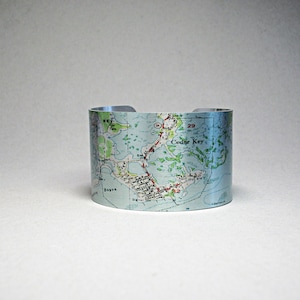 May include: A silver metal cuff bracelet with a map of Cedar Key, Florida printed on it. The map shows roads, waterways, and the text "Cedar Key".