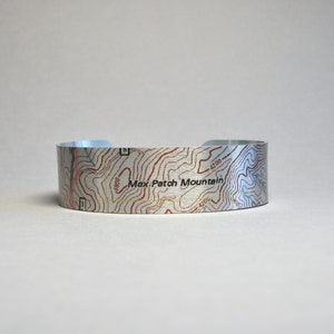 Peut inclure: Un bracelet en métal argenté avec un motif de carte topographique comportant des lignes de niveau et le texte "Max Patch Mountain".