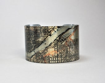 May include: Wide cuff bracelet with a map design. The bracelet features a silver-colored interior and a wrap-around design with a detailed map print in shades of black, gray, and orange. The cuff is approximately 2 inches wide.
