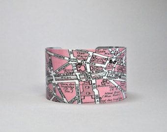 May include: A wide cuff bracelet featuring a printed map of Paris, France. The design displays pink street blocks, white roads, and black text labels for landmarks such as the Grand Palais, Seine, and Hotel des Invalides on a curved metal band.