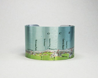 Puede incluir: Una pulsera de puño ancha con un diseño de mapa. La pulsera presenta un fondo azul claro con un mapa detallado de una zona costera, incluyendo nombres de lugares como Cannon Beach y Tolovana Park. El borde inferior tiene detalles verdes y blancos.