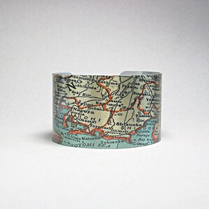 May include: A metal cuff bracelet with a vintage map of Japan printed on it. The map shows the islands of Honshu, Shikoku, and Kyushu, with major cities and geographic features labeled. The map is in shades of blue, green, and brown, with red lines indicating roads.