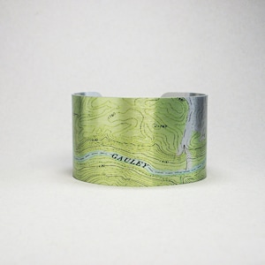 May include: A silver metal cuff bracelet with a topographic map design. The map features green and brown contour lines and the word "GAULEY" printed in black.