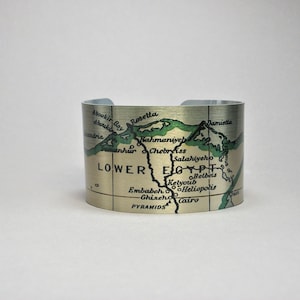 May include: A gold metal cuff bracelet with a map of Lower Egypt printed on it. The map shows the Nile River, major cities, and the pyramids. The map is in green and black on a gold background.