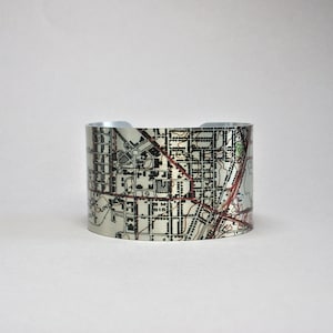 May include: A wide, silver cuff bracelet with a map design in grey, black, and red. The bracelet has a smooth, metallic finish and a rectangular shape. The cuff measures approximately 5 cm in width.
