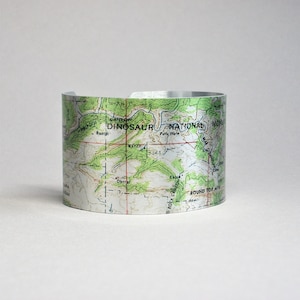 May include: A silver metal cuff bracelet with a map of Dinosaur National Monument printed on it. The map shows the location of Winepool Canyon, Ranch, Pats Hole, Corral, Cabin, and O Rock Canyon.
