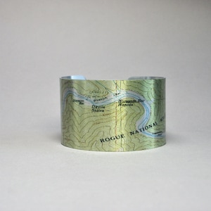 May include: A silver metal cuff bracelet with a map of the Rogue National Wilderness in Oregon. The map shows the location of Blossom Bar Rapids, Devil's Stairs, and other points of interest.