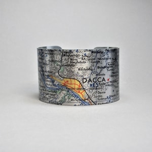 May include: A silver metal cuff bracelet with a map of Dhaka, Bangladesh printed on it. The map shows the city's streets, rivers, and landmarks. The word "DACCA" is printed in red on the map.