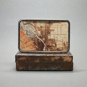 May include: A silver belt buckle with a map of a city on it. The map is in shades of brown and white and shows a river running through the city. The buckle is sitting on a rusty metal box.