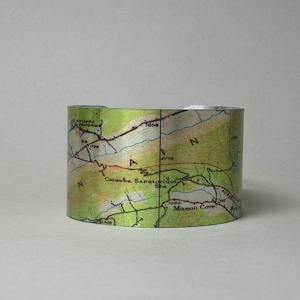 May include: A cylindrical metal cuff bracelet with a vintage map design. The map shows green, tan, and white topographical details, with place names and contour lines. The bracelet has a metallic sheen and a smooth finish.