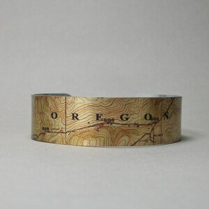 May include: A wide, open cuff bracelet featuring a topographic map design. The map is printed in shades of brown and tan, with the word "OREGON" visible. The bracelet is a neutral accessory.