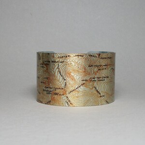 May include: A gold metal cuff bracelet with a topographic map design of the Appalachian Mountains. The map features contour lines and labels for various peaks and trails.