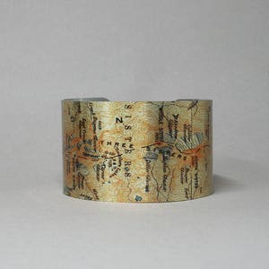 Puede incluir: Un brazalete de metal con un mapa de la Wilderness de las Tres Hermanas en Oreg&oacute;n. El mapa est&aacute; impreso en marr&oacute;n y dorado sobre un fondo plateado. El texto del mapa incluye "Three Sisters", "Middle Sister", "South Sister" y "Glacier".