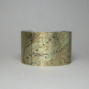 Peut inclure: Un large bracelet manchette avec un motif de carte topographique. La carte est imprimée sur un fond clair avec des lignes de contour marron et du texte, dont "Mount Madison" et "Max Patch Mtn". Le bracelet est de forme incurvée.